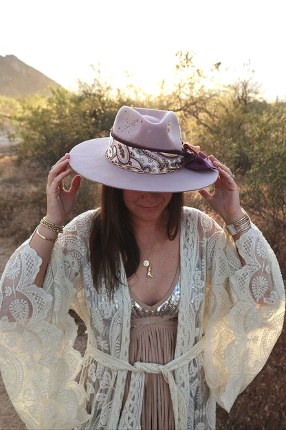 A woman outdoors wearing a wide-brimmed lavender hat with a patterned band, a cream lace cardigan, and a beige dress, with hills and shrubs in the background.